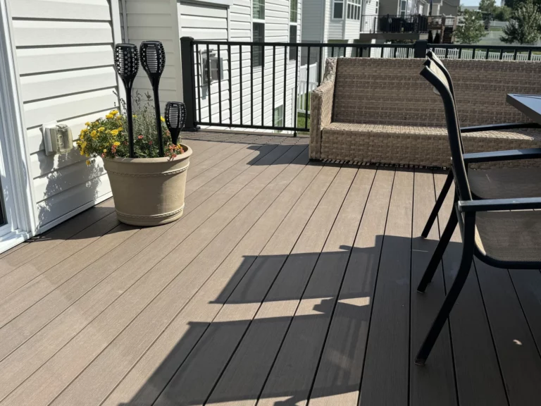 A spacious outdoor deck featuring a wicker sofa, a dining table with black chairs, and a decorative pot filled with bright flowers and torch planters under a sunny sky.