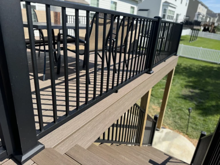 A view of a wooden deck with black railings and chairs, showing a staircase leading down to a grassy area.