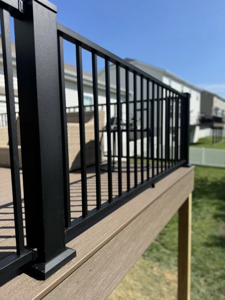 Close-up view of a black railing on a wooden deck, with a clear blue sky and neighboring houses in the background.