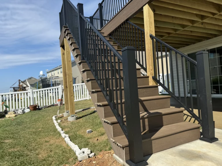 A staircase leading up to a deck, featuring dark metal railings and wooden steps, set in a backyard with a grassy area and a white picket fence in the background.