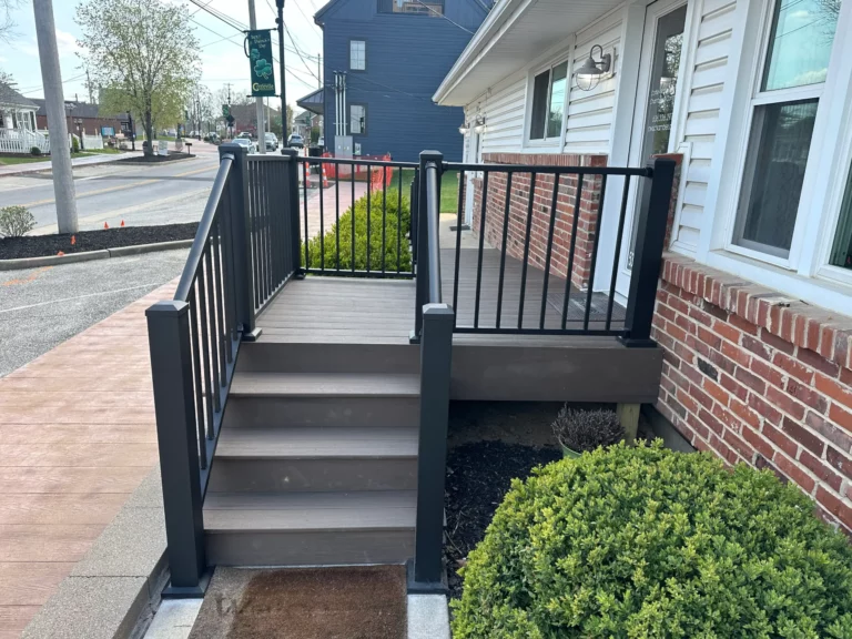 A view of a porch with black railings and brown steps leading to a house, surrounded by green bushes and a neighborhood street in the background.