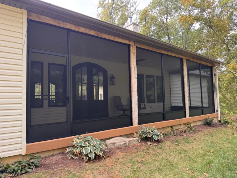 A modern screened porch with large black screens and wooden accents, featuring a comfortable chair inside and surrounded by greenery.