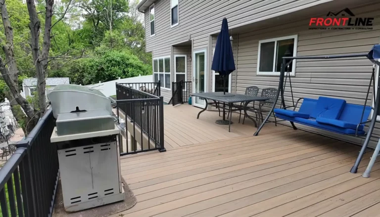 A spacious wooden deck with a stainless steel grill on one side, a dining table with chairs, and a blue swing chair, surrounded by lush green trees.