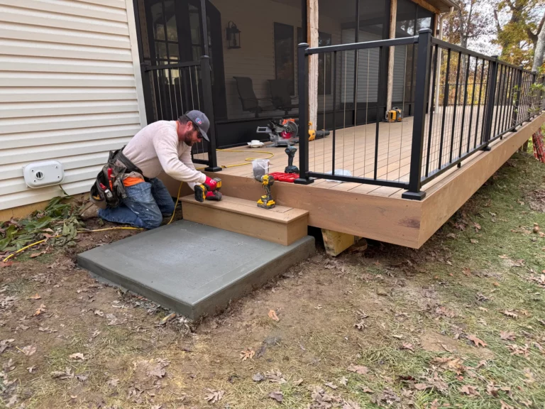 A man is working on a wooden deck, installing a step near a concrete base. He is kneeling on the ground with tools around him, wearing a gray hat and a work apron. The deck has black railings and is in a yard with fallen leaves.