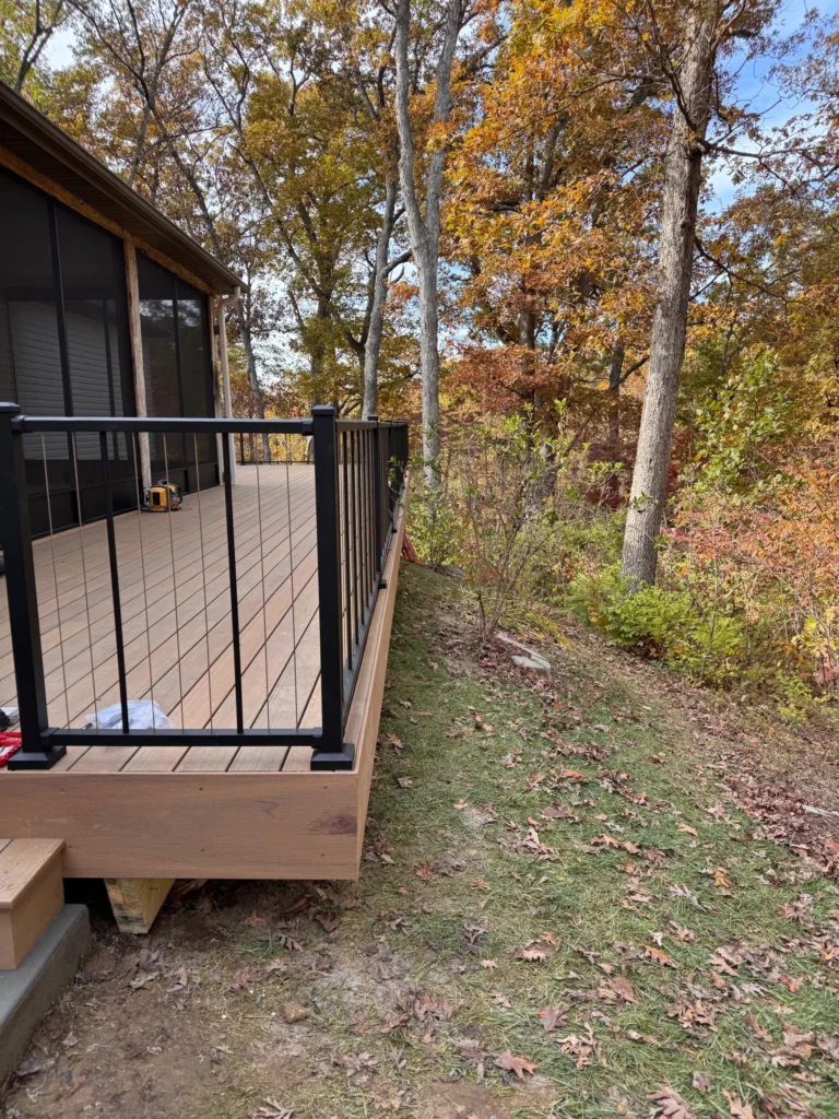 A modern deck viewed from the side, featuring a black railing, surrounded by trees with autumn foliage. The floor is made of wood and is partially covered with fallen leaves.