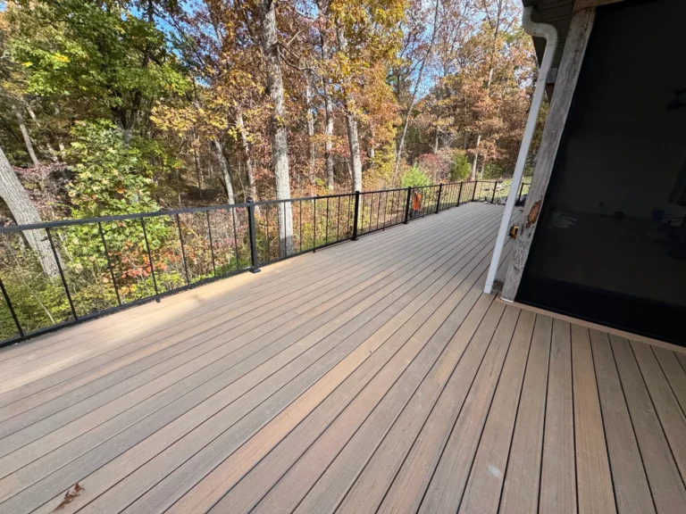 A spacious wooden deck with black railings overlooks a vibrant forest in autumn, featuring colorful leaves on the trees and a clear blue sky.
