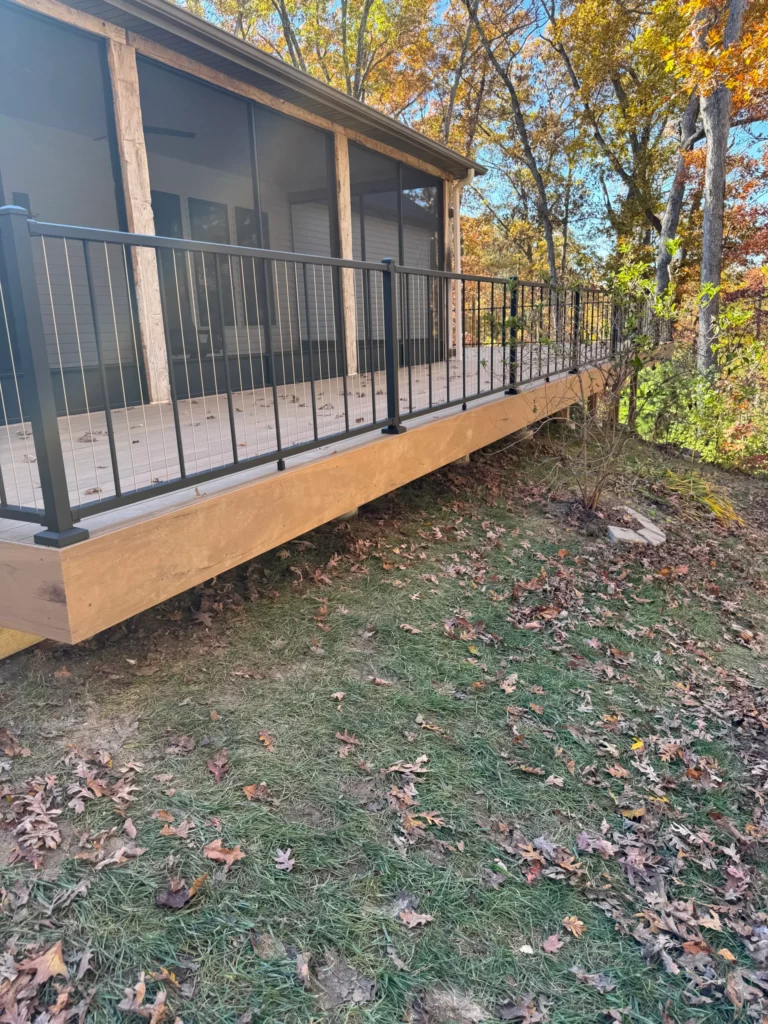 A view of a wooden deck with a black railing, overlooking a yard covered with fallen leaves. Trees with colorful autumn foliage surround the area.