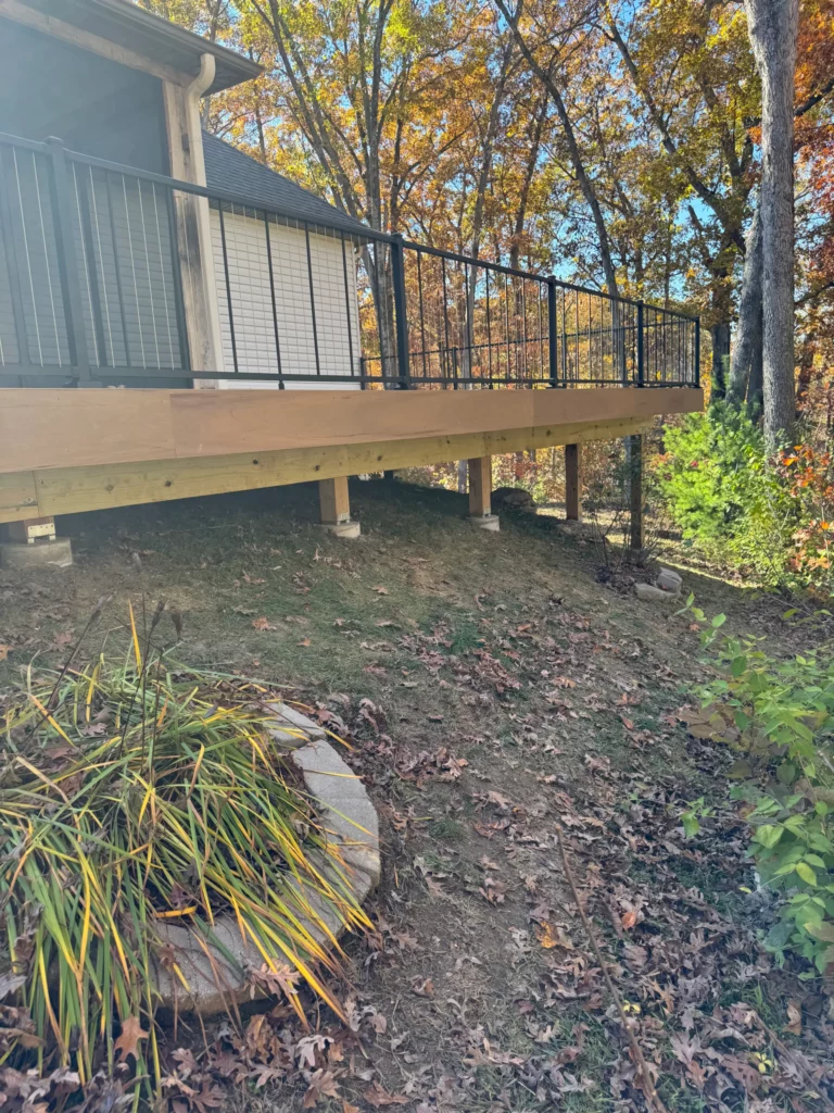 A wooden deck with a black railing overlooks a landscaped area filled with autumn leaves and shrubs, surrounded by trees with colorful fall foliage.